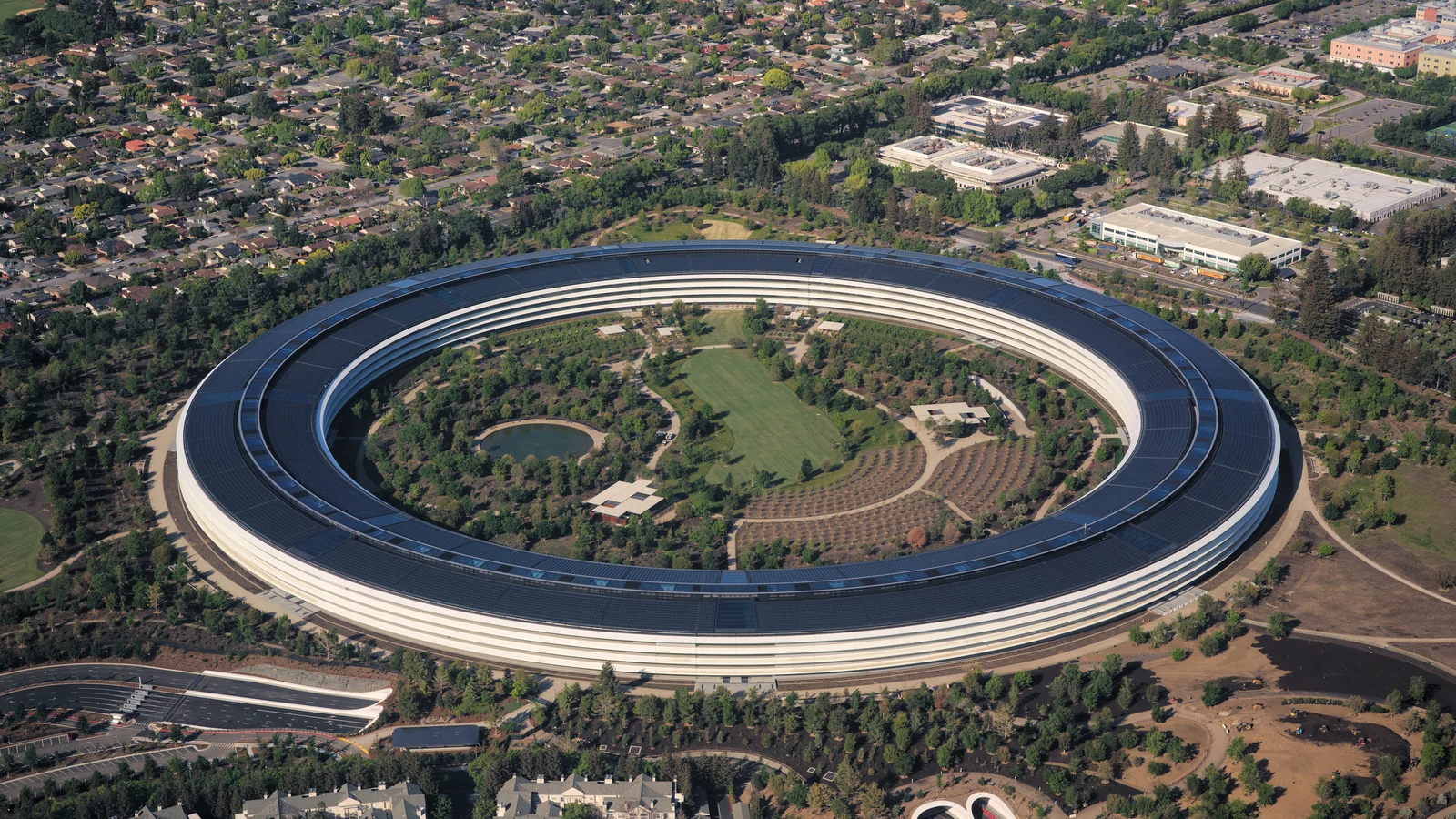 An aerial view shows a large elliptical, ring-shaped multi-story building with layered white exterior bands and a dark roof covered in rectangular panels.